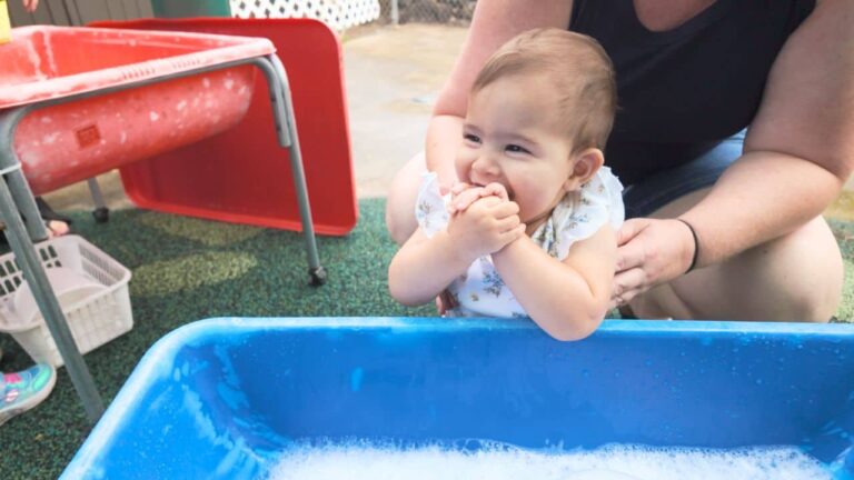 Baby in a floral outfit chews a cloth beside a blue water tub while an adult supports her; red play equipment fills the outdoor yard.