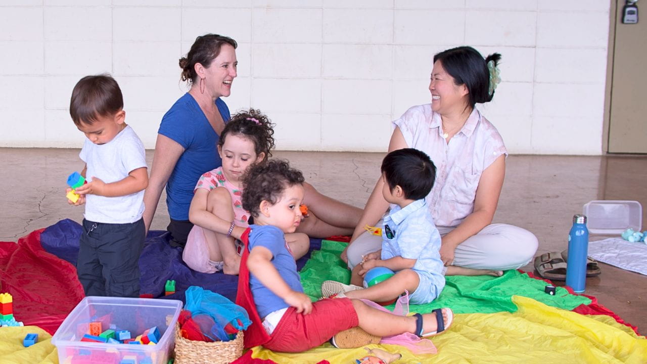 Two women and four young children sit on colorful blankets indoors, smiling and playing with toys in a warm, joyful group scene.