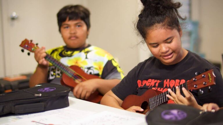 Two young musicians practice ukulele indoors: a focused girl strums in front while a boy plays behind her, surrounded by instrument cases.