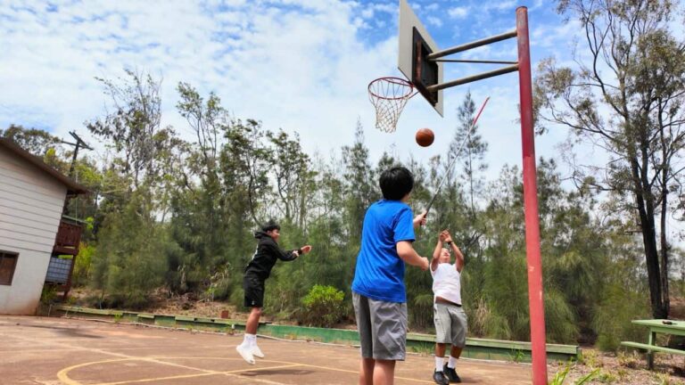Three teens play basketball on an outdoor court; a blue-shirted player taps the hoop with a walking aid as others jump and watch among trees.