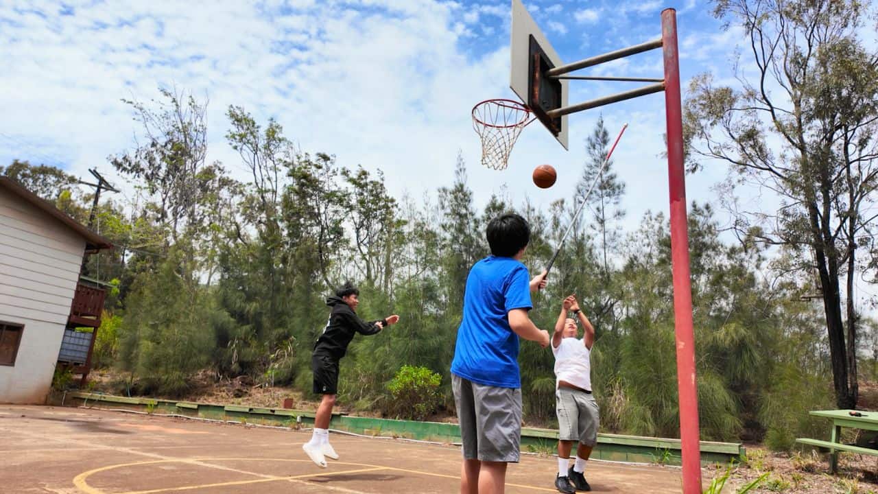 Three teens play basketball on an outdoor court; a blue-shirted player taps the hoop with a walking aid as others jump and watch among trees.