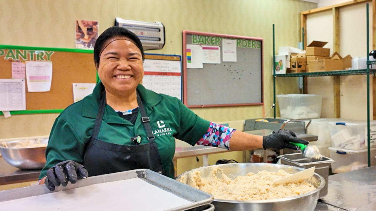 Smiling bakery worker in green uniform and apron mixes dough in a large bowl inside a busy commercial kitchen, creating a warm, cheerful mood.